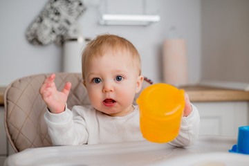 baby is sitting in a high chair, holding an orange. a baby in a high chair drinks water from an orange drinking bowl