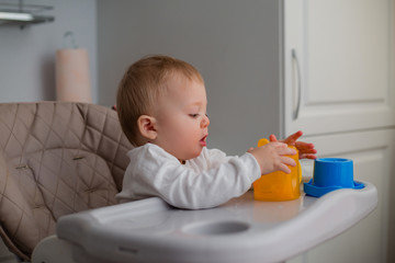 baby is sitting in a high chair, holding an orange. a baby in a high chair drinks water from an orange drinking bowl