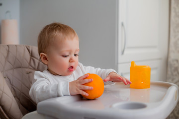 baby is sitting in a high chair, holding an orange. a baby in a high chair drinks water from an orange drinking bowl