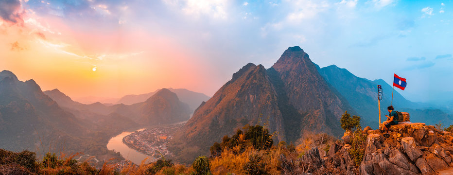 Sunset panoramic view of couple of trekkers sitting on a rock on top of Nong Khiaw View Point with beautiful mountain and Nam Ou river in background, Luang Prabang Province, Northern Laos.