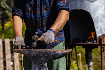 A close up shot on the muscular arms of a blacksmith at work using a hammer and anvil to shape iron, with blurry furnace in background, copy space to right