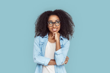 Freestyle. African girl in glasses standing isolated on gray touching chin looking aside playful