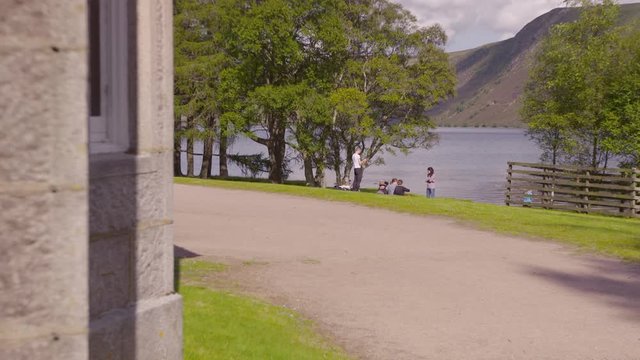 Wide Low Angle Still Shot From Queen Victoria Lodge House Of People Relaxing On Green Grass Next To Lush Green Trees Near River Muick ( Loch Muick), And Horizon Hilly Landscape Against The Sky