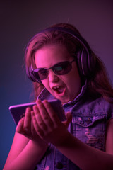 Girl in denim vest with big headphones and sunglasses looks at the contents on her phone and grins