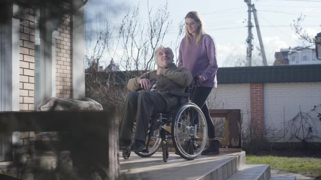 Long shot of adult female invalid tender talking with old Caucasian man and rolling wheelchair away. Smiling nurse taking care of elderly disabled retiree. Assistance, help, nursing.