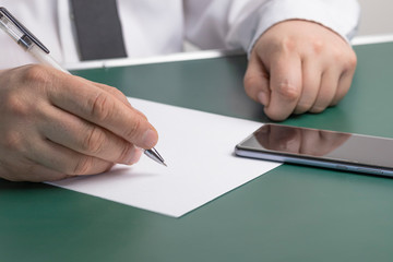 the businessman writes on a sheet of white paper next to the phone. Wood background. Dressed in a shirt and tie. Close-up.