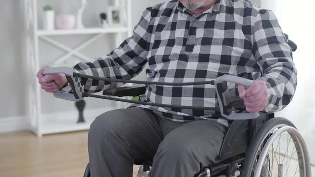 Old unrecognizable Caucasian man in wheelchair stretching elastic band. Elderly male disabled person exercising indoors. Physical therapy, health care, disability.