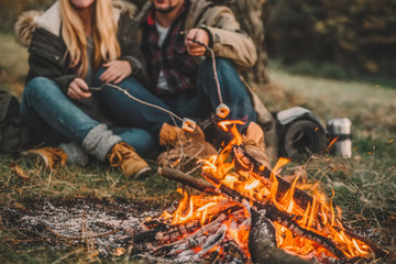 Traveler couple camping and roasting marshmallows over the fire in the forest after a hard day. Concept of trekking, adventure and seasonal vacation.