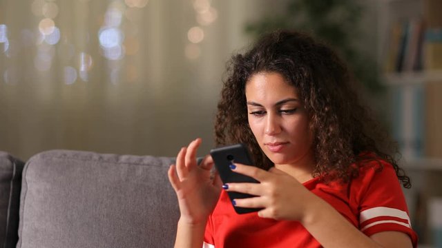 Serious arab girl in red checking smart phone sitting on a couch in the living room at home in the night