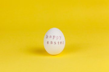 One white chicken egg with words Happy Easter written with help of stamps and pink metallic paint on yellow background, copy space. DIY, festive concept. Side view. Horizontal