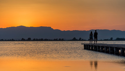 Naklejka na ściany i meble A young couple watches the sunset on a pier at the mouth of the Ebro river, Trabucador beach, in the Mediterranean Sea, in the province of Tarragona (Spain).