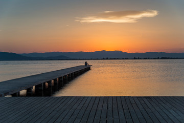 Fototapeta premium A young couple watches the sunset on a pier at the mouth of the Ebro river, Trabucador beach, in the Mediterranean Sea, in the province of Tarragona (Spain).