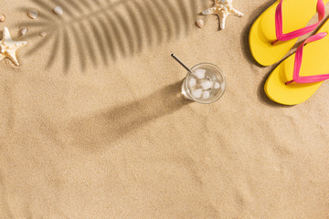 Fototapeta na wymiar Summer fashion, summer outfit on sand background. Yellow flip flops, glass of water and seashells. Flat lay, top view. Harsh light with shadows