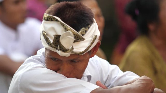 Close up view of Balinese man in traditional clothing as he waits for Hindu temple ceremony. Older man with weathered brown skin wipes face on arm and looks into camera smiling.