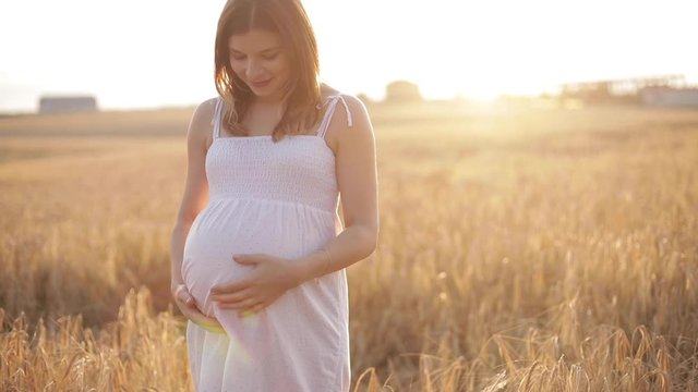 Close-up 4K video of an incognito pregnant woman in white polka dot dress touching and patting her belly and making heart shape with her hands. She is standing in sunlit meadow.