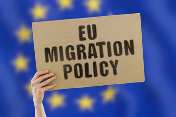 The phrase " EU migration policy " on a banner in men's hand. Human holds a cardboard with an inscription. Travel policy. Tourism. European Union flag. Trip. Rules. Immigrants. Refugee. Cross border