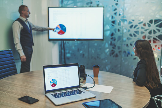 A Business Meeting In A Board Room With Selective Focus On A Laptop And Gadgets In The Foreground And Two Partners In The Background: Man Entrepreneur Is Showing On The Diagram On The Screen Of Plasma