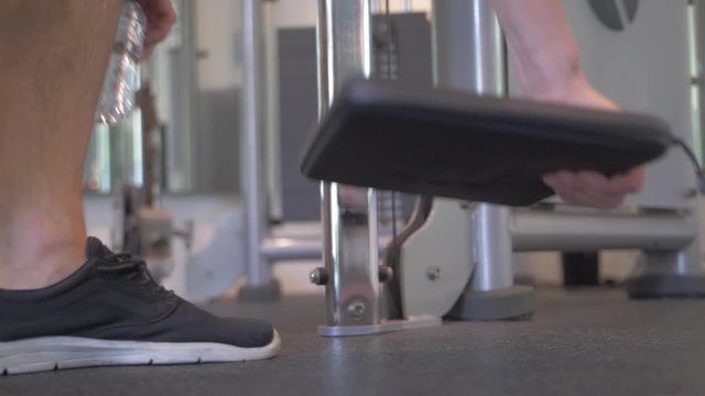 Detail shot of a white, athletic man putting down a black laptop case and a water bottle on the floor of a gym before starting his workout and exercise. Slow motion.