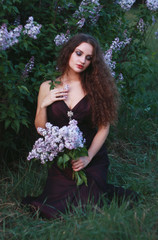A woman dressed in a beautiful purple dress on a background of lilac bushes with a bouquet of lilac in the hands of spring.