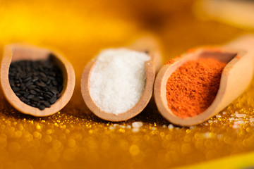 small wooden spoons with black sesame, salt and papic on a yellow shiny background