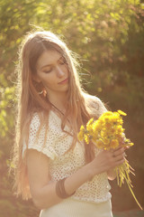 A woman dressed in a white knitted blouse and beige shorts in nature with a bouquet of dandelions.