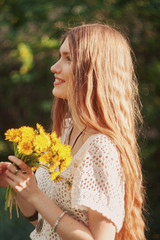 A woman dressed in a white knitted blouse and beige shorts in nature with a bouquet of dandelions.