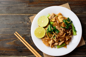 Stir-fried soy sauce rice noodles with pork on white plate and chopsticks on wooden  background, Thai food (Pad See Ew), Top view