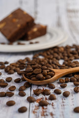 Coffee beans on wooden spoon and chocolate brownies on a white wood table.