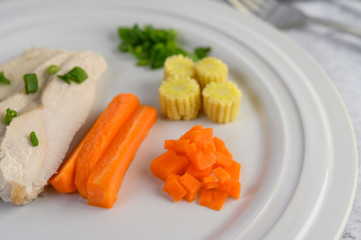 Steamed chicken breast on a white plate with spring onions, baby corn, and chopped carrots.