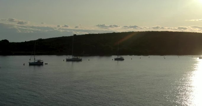 A video of a bunch of anchored sail boats in between the two islands. The sun is shinning directly into the camera, the drone is moving sideways. Video was filmed during sunrise.