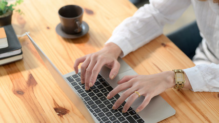 Cropped shot of a woman typing on laptop on wooden desk in simple workplace