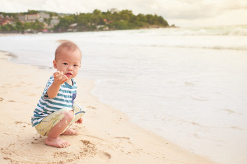 Cute little Asian toddler baby boy child on beach with dirty hands covered with wet sand. Family travel, water outdoor activity on summer beach vacation with children, Sensory play with sand concept