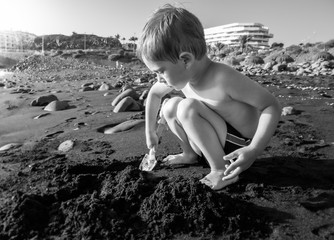 Black and white portrait of little toddler boy digging sand on the ocean beach at sunset