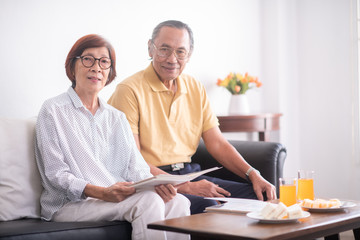 Obraz premium Asian old couple sitting in living room having snack and looking at photo album with happy feeling.healthy and positive retired people concept