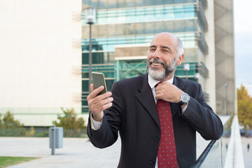 Smiling mature man holding smartphone and adjusting tie. Cheerful bearded preparing himself before important video call while standing near office building. Video call, technology concept