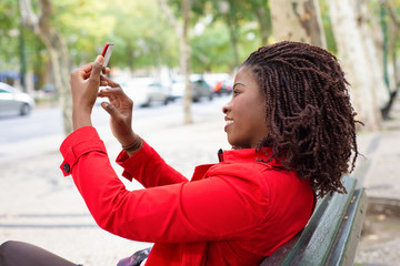 Side view of beautiful woman taking selfie with smartphone. Cheerful young lady with dreadlocks holding smartphone in outstretched arms while sitting on bench in autumn park. Self portrait concept
