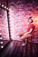 Girl in red shirt on a tall chair in front of a lighted mirror in a dark room. Model posing for a...