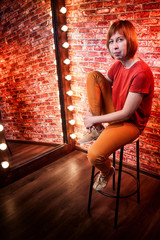 Girl in red shirt on a tall chair in front of a lighted mirror in a dark room. Model posing for a...