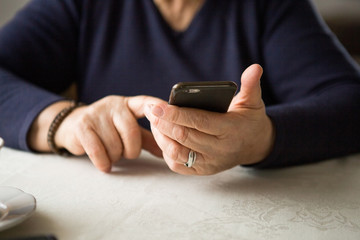 Retired woman is using mobile phone,  at cozy home kitchen. Grandmother has coffee break in cooking. Senior person is looking for recipe, communication online in the internet. Authentic moment.