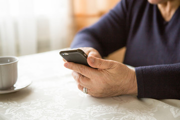 Retired woman is using mobile phone,  at cozy home kitchen. Grandmother has coffee break in cooking. Senior person is looking for recipe, communication online in the internet. Authentic moment.