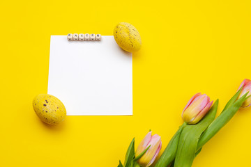 Naklejka na ściany i meble Easter background with colorful eggs, pink tulips and greeting card over yellow background. Top view with copy space.