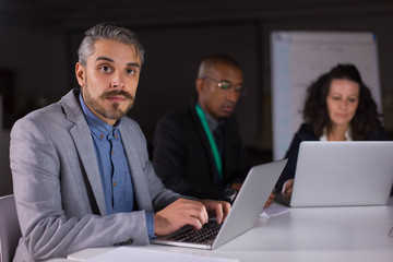 Fototapeta premium Concentrated manager sitting at table and looking at camera. Focused man working with laptop in office at night. Business, working late concept