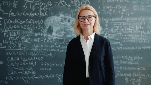 Slow motion of friendly teacher wearing suit and glasses smiling looking at camera standing alone in classroom, blackboard with scientific formulas is in background.