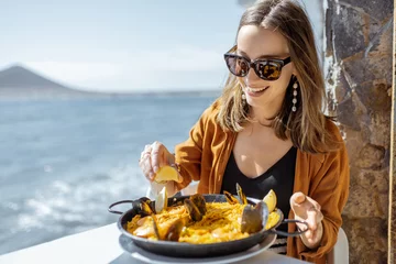 Gordijnen Vrouw eet paella, traditioneel Spaans gerecht, zittend op het terras van het restaurant in de buurt van de oceaan. Concept van zeevruchten en goede zomervakanties © rh2010