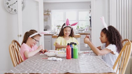 Group Of Girls Wearing Bunny Ears Sitting At Table Decorating Eggs For Easter At Home