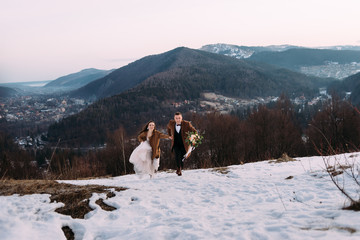 stylish couple of newlyweds running in the snow, men's coat brown, bouquet for the wedding. Holidays for honeymooners at a ski resort