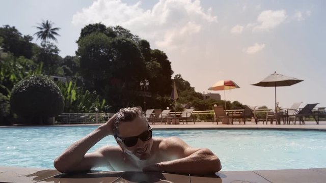 Handsome man with braces in sunglasses enjoying water in swimming pool. Portrait of relaxed smiling guy with tanned skin combing wet hair with hand