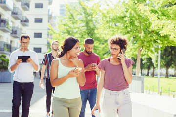 Obraz premium Group of gadget addicted people walking outside. Young men and women talking on cellphones and using smartphones and tablet. Devices addiction concept