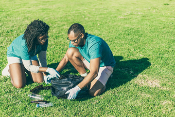 Volunteers picking up bottles outside. Black woman and Latin man wearing uniforms and protective...