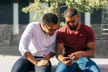 Excited male friends studying mobile app features on phone. Two men in casual sitting on parapet outside, using one smartphone and staring at screen. Mobile app concept